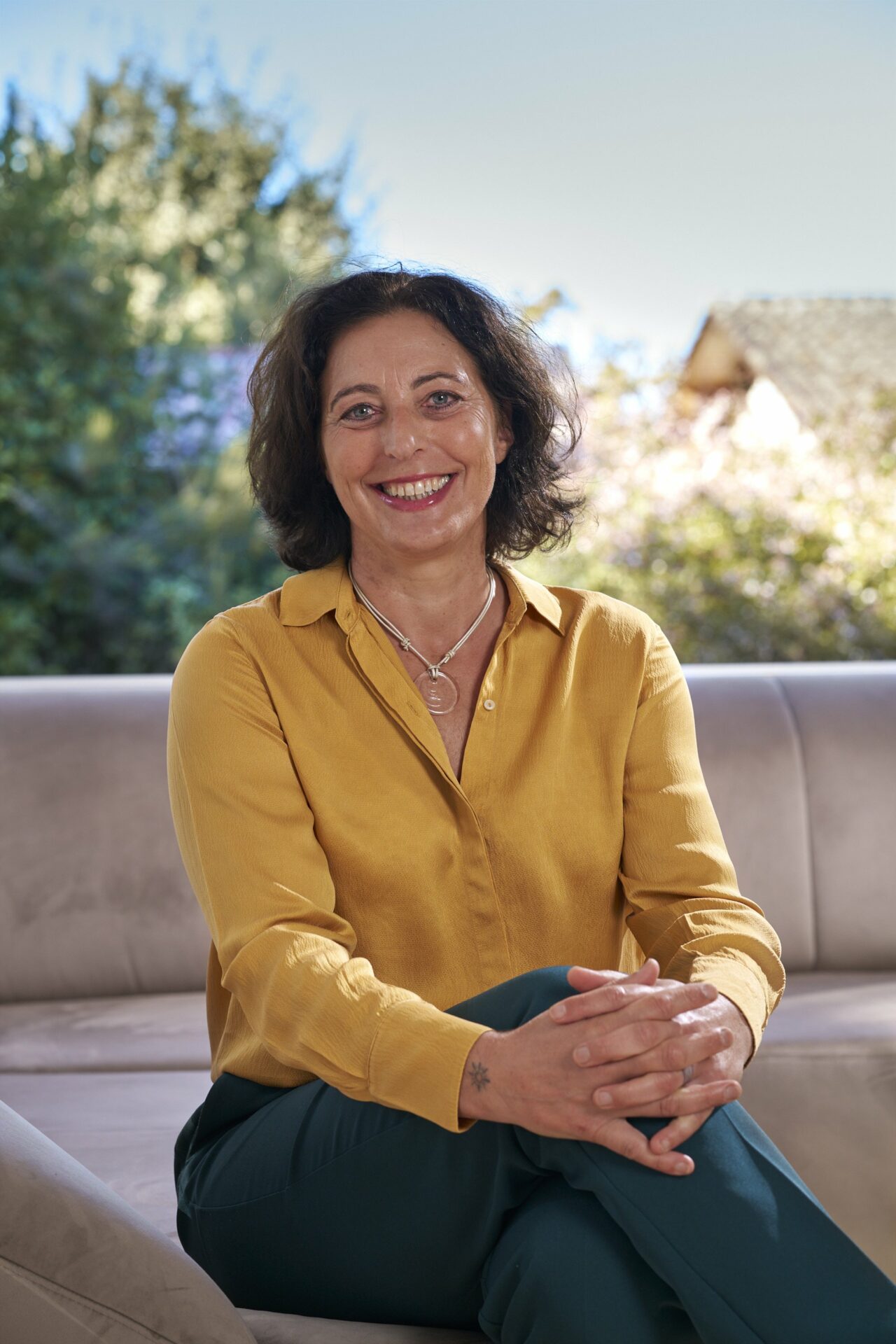 Melina Jobin assise sur un canapé en espace de séance, souriante, portant une chemise jaune, avec un jardin en arrière-plan.